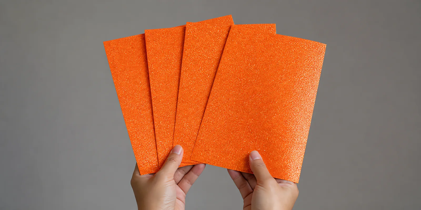 Manos sosteniendo varias hojas de papel con glitter naranja soleado abiertas en abanico