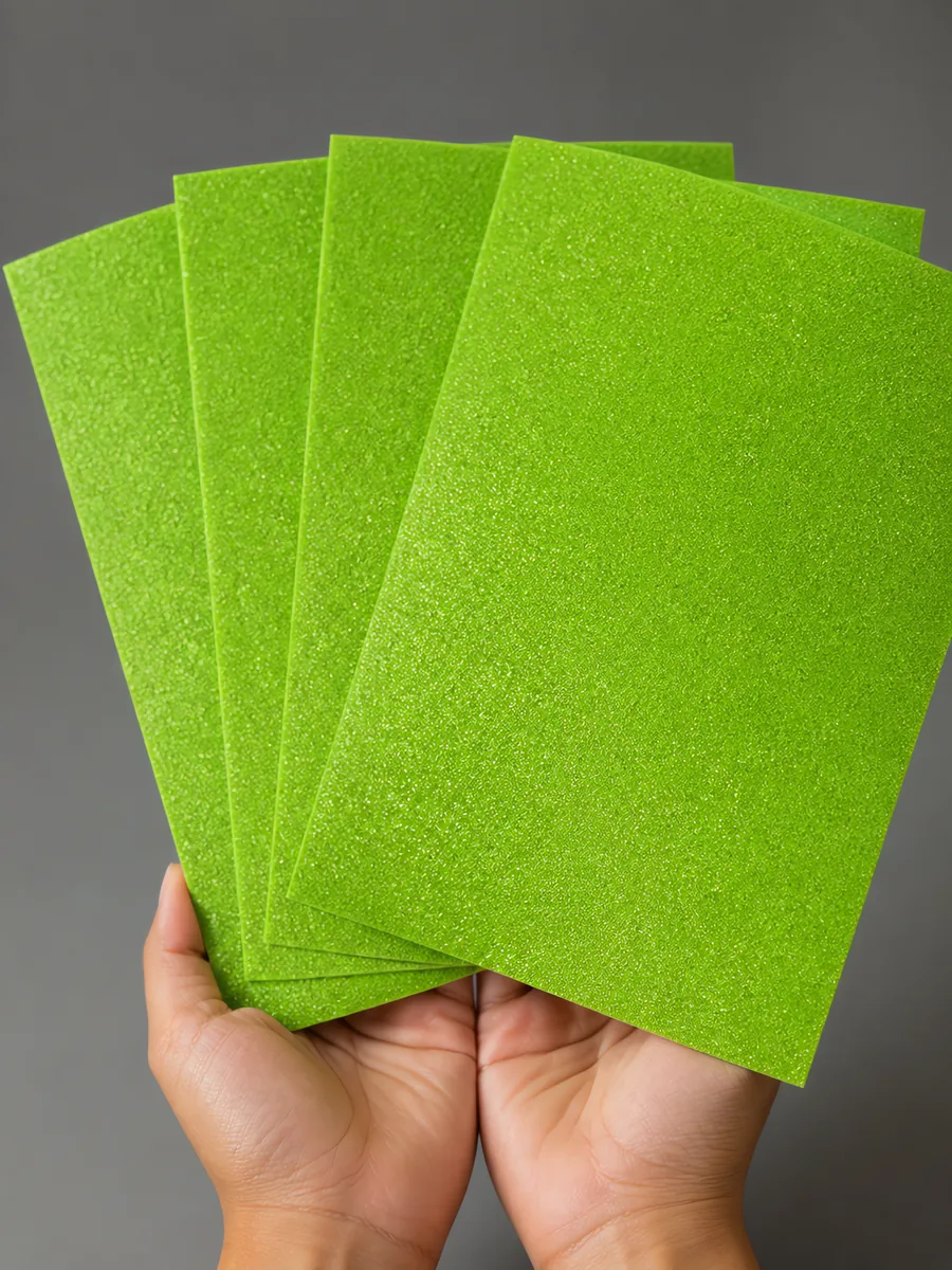 Manos sosteniendo varias hojas de papel con glitter verde jardin reluciente abiertas en abanico