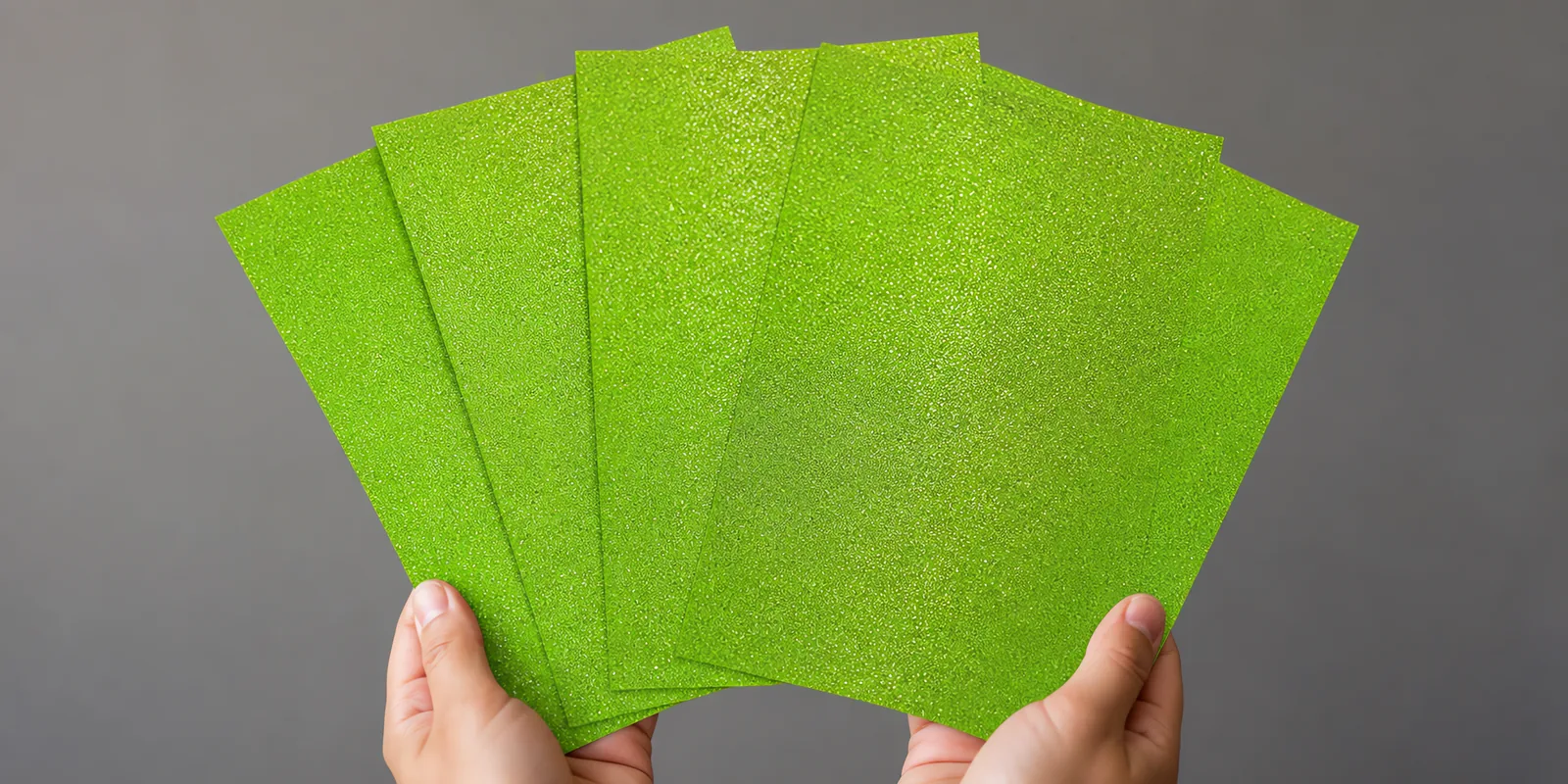 Manos sosteniendo varias hojas de papel con glitter verde jardin reluciente abiertas en abanico