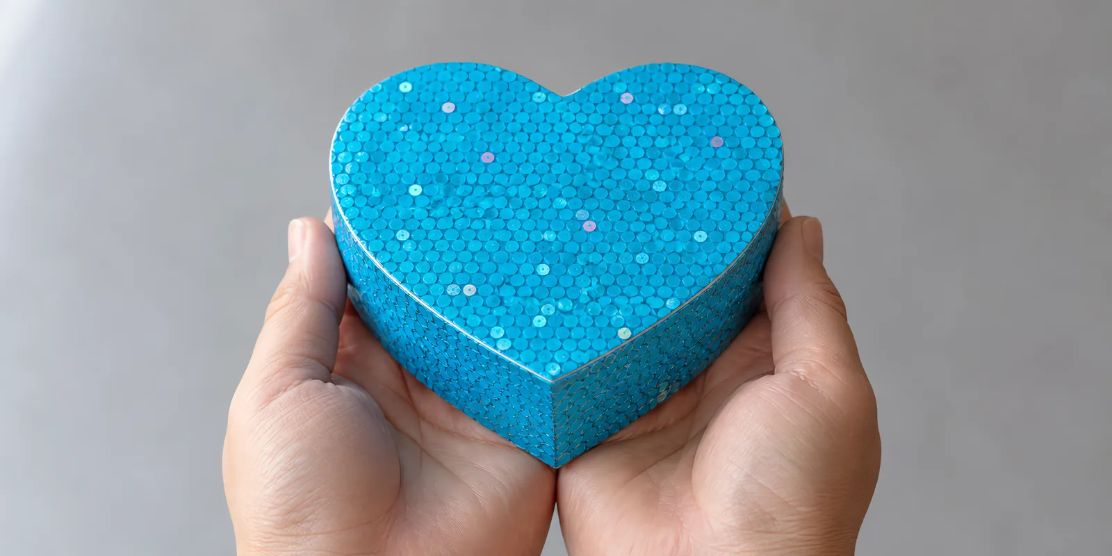 Manos sosteniendo una cajita de corazón de papel con glitter en escamas, en el tono azul katy