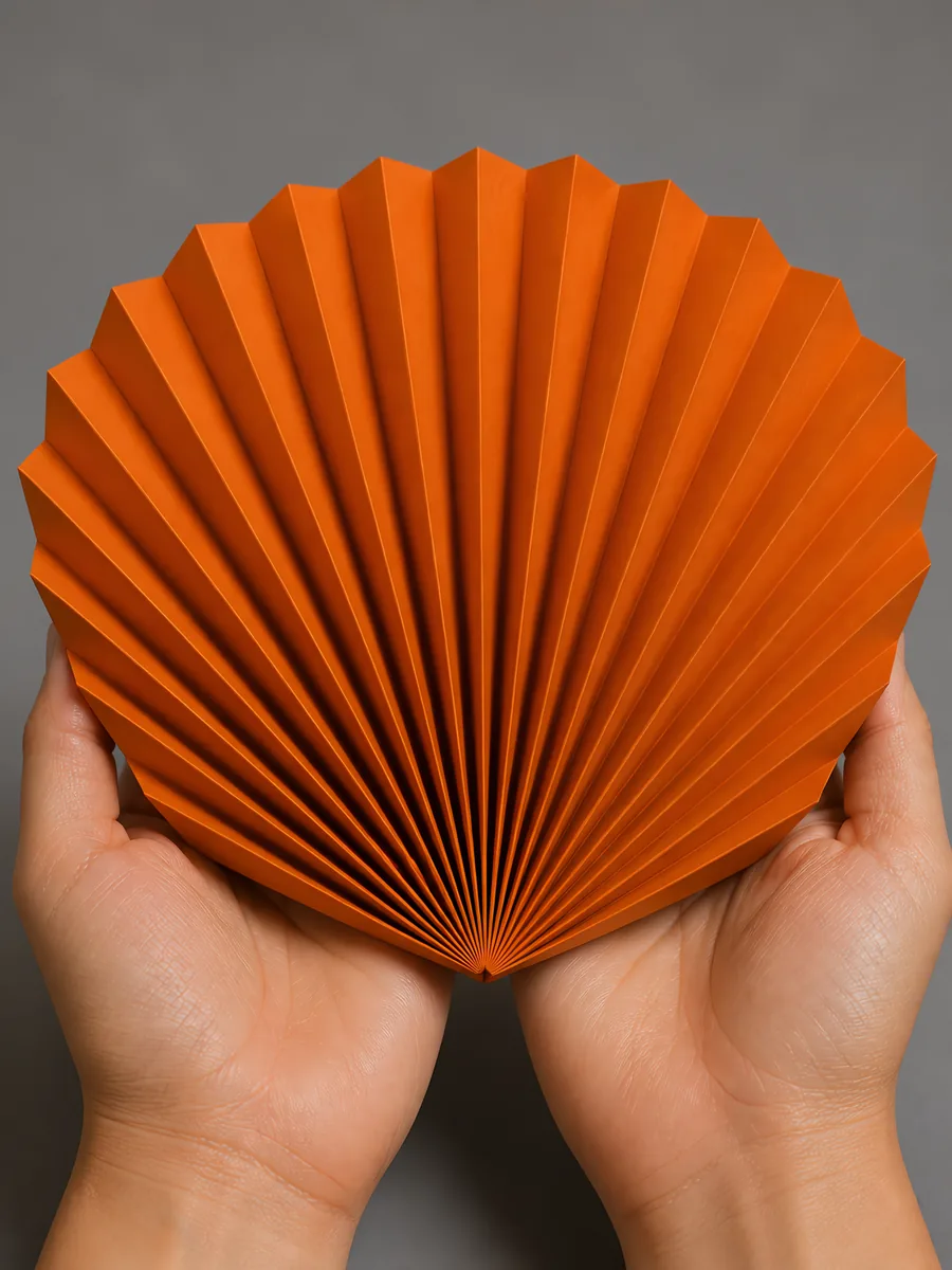 Manos sosteniendo un abanico de papel plegado en acordeón, en naranja caramelo
