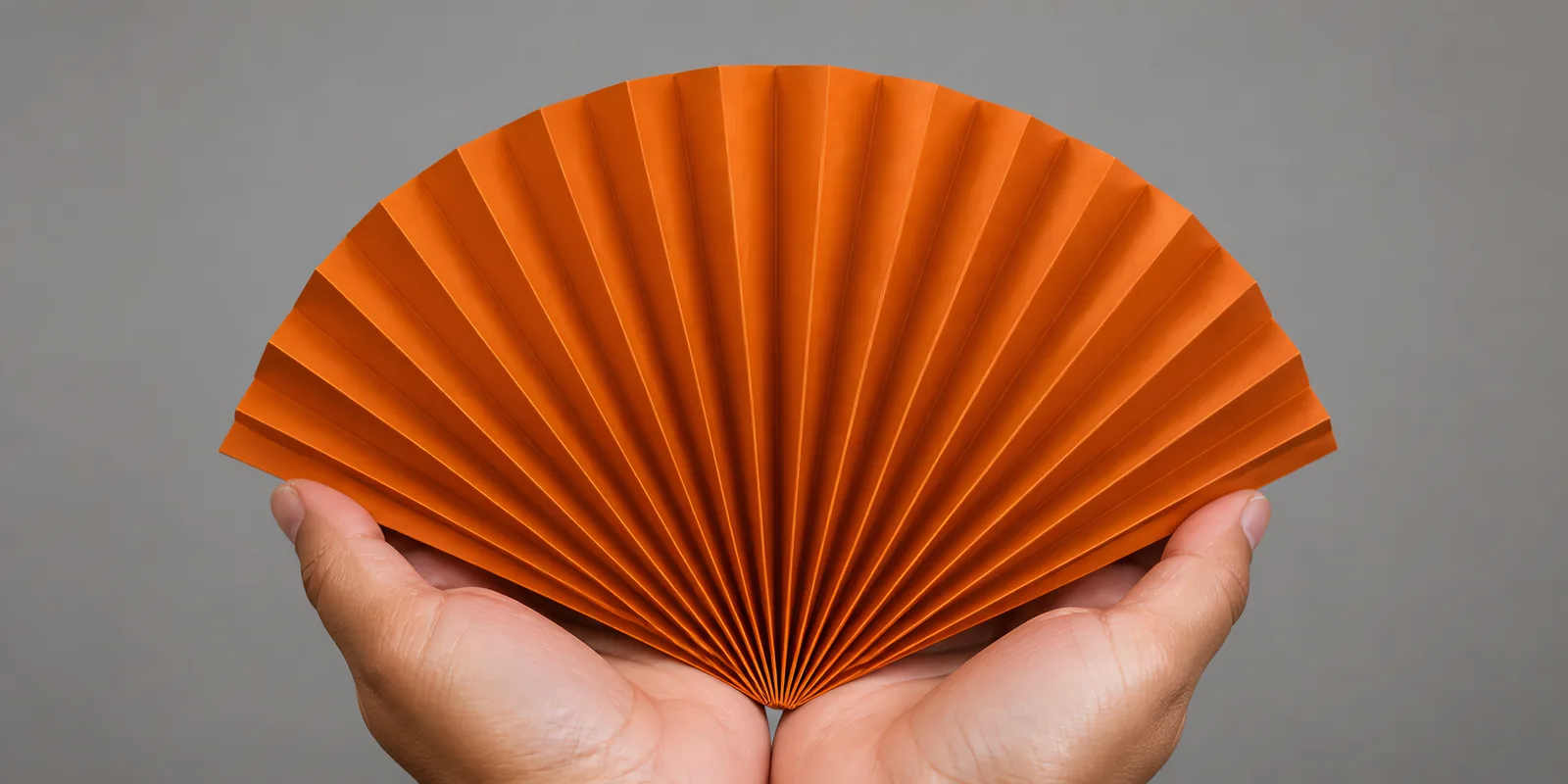 Manos sosteniendo un abanico de papel plegado en acordeón, en naranja caramelo