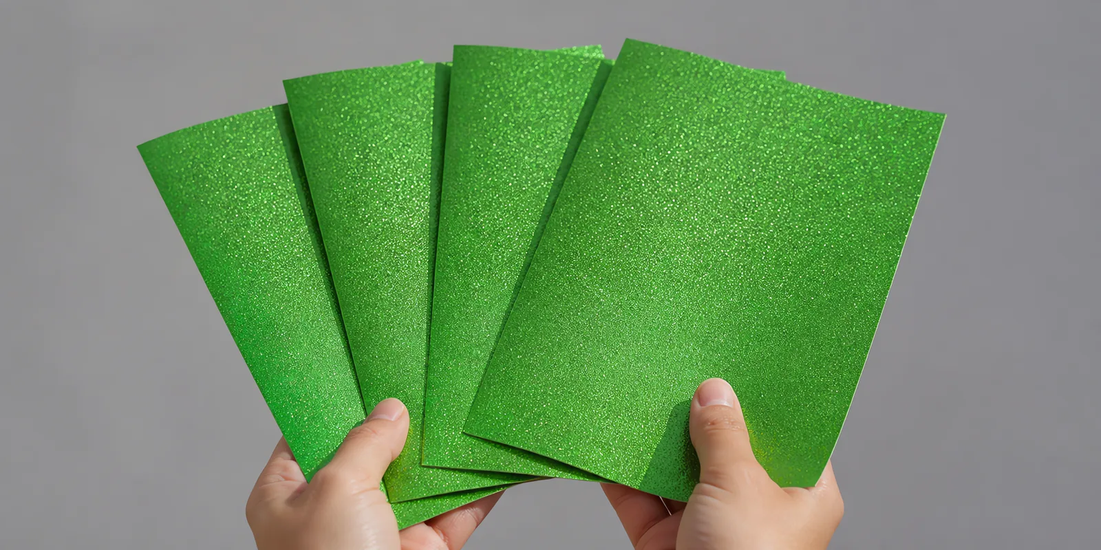 Manos sosteniendo varias hojas de papel con glitter verde bosque encantado abiertas en abanico