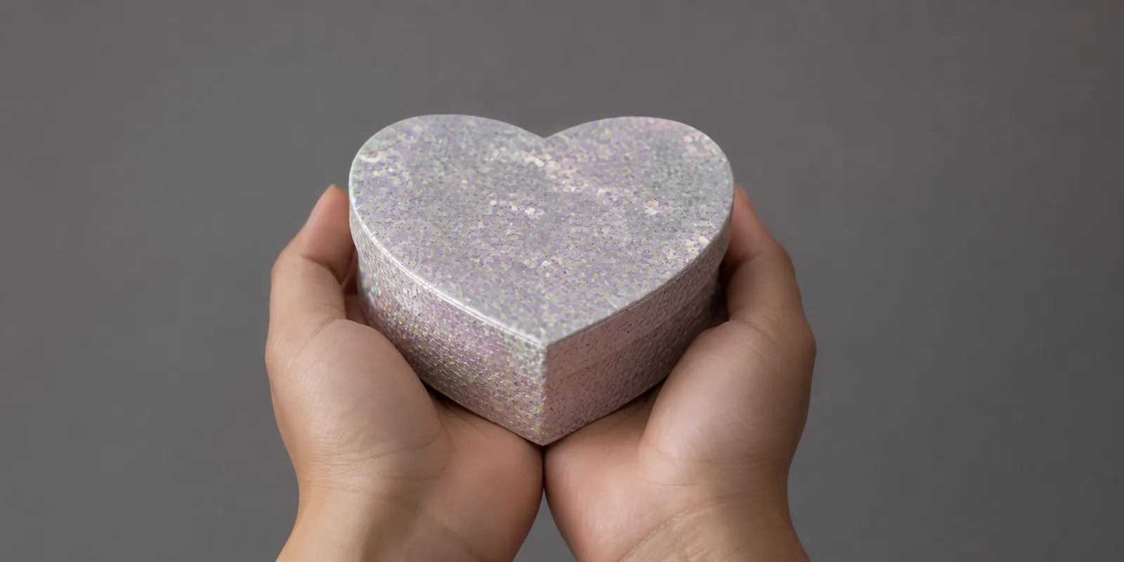 Manos sosteniendo una cajita de corazón de papel con glitter en escamas, en el tono iridiscentes dua
