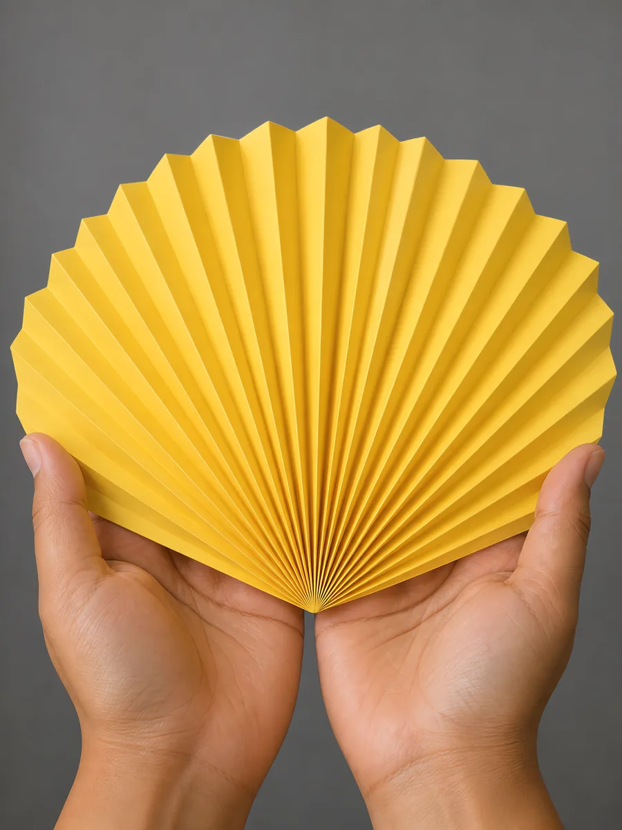Manos sosteniendo un abanico de papel plegado en acordeón, en amarillo girasol
