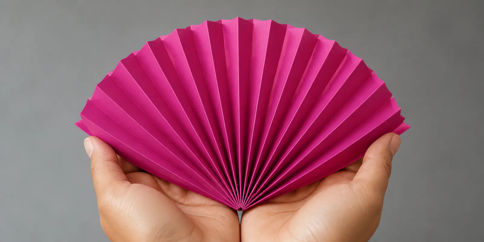 Manos sosteniendo un abanico de papel plegado en acordeón, en fucsia chic