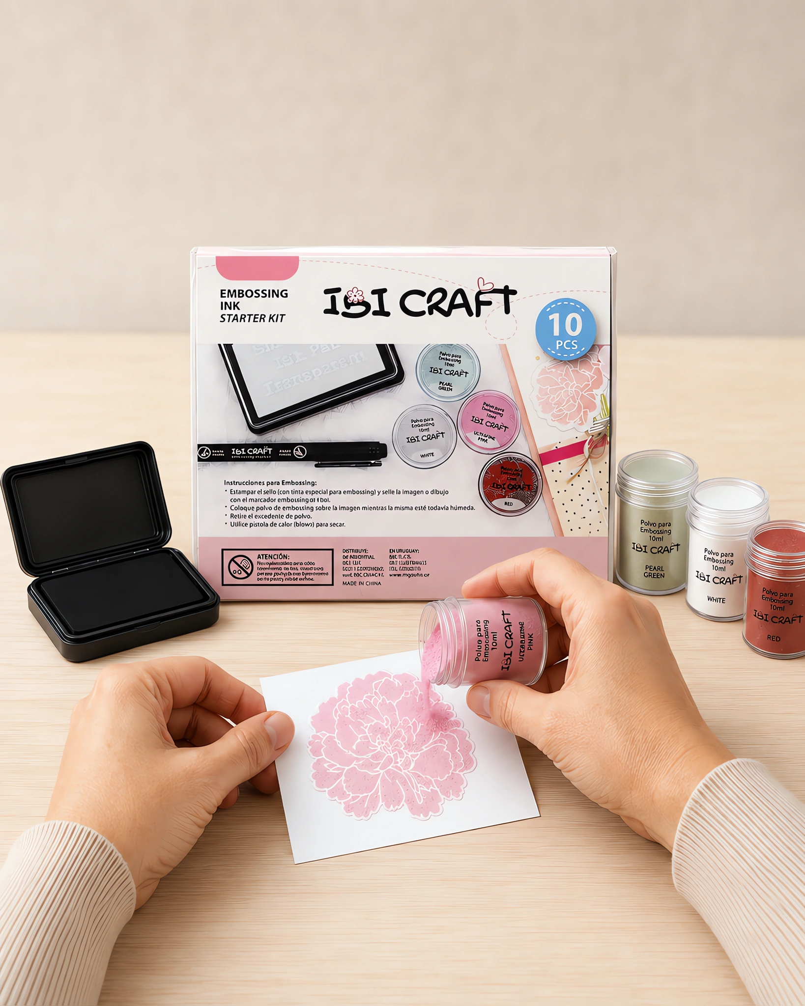 Manos espolvoreando polvo de embossing sobre una imagen recién entintada, con la caja del kit al lado